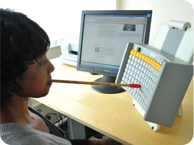 Person typing with a mouth stick. The keyboard is positioned vertically
      on a table in front of their face, and sits next to the computer monitor.