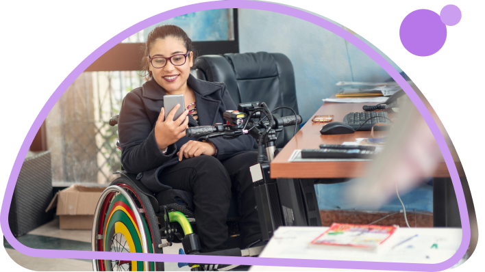 A woman with glasses and hair pulled back smiling, looking at her phone, sitting in a colorful wheelchair